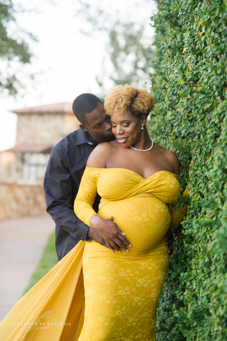 Mommy smiling while daddy to be kisses her against a vine wall at Bella Collina 